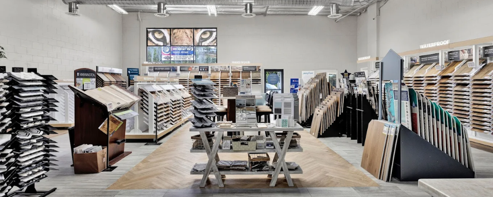 A well-lit flooring store with displays of various luxury vinyl wood-look and tile samples arranged on shelves and racks, featuring a large screen displaying tiger eyes and nature scenes on the back wall.